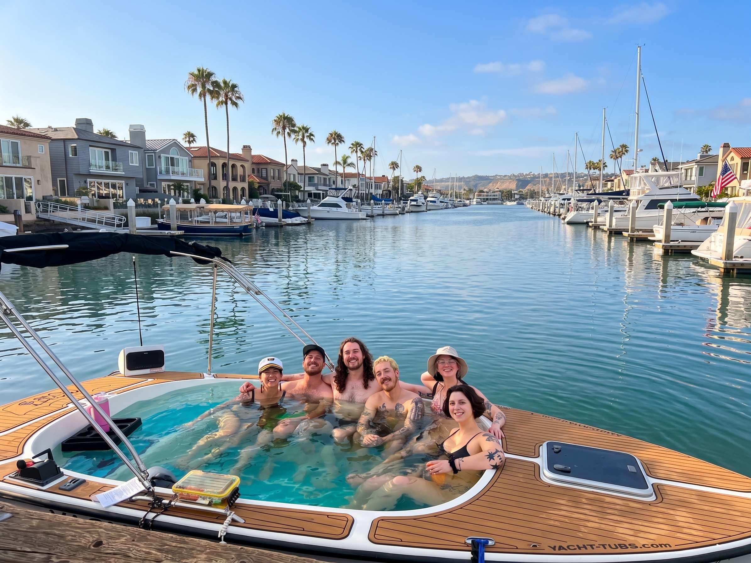Six guests soaking in a hot tub boat on Newport Harbor, with waterfront homes, palm trees and a docked Duffy boat behind them